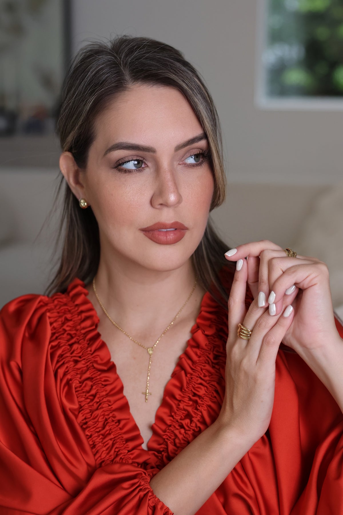 A woman with styled brown hair is wearing a vibrant red satin blouse with ruffled trim and gold jewelry, including a delicate gold necklace and rings, set against a softly blurred indoor background.